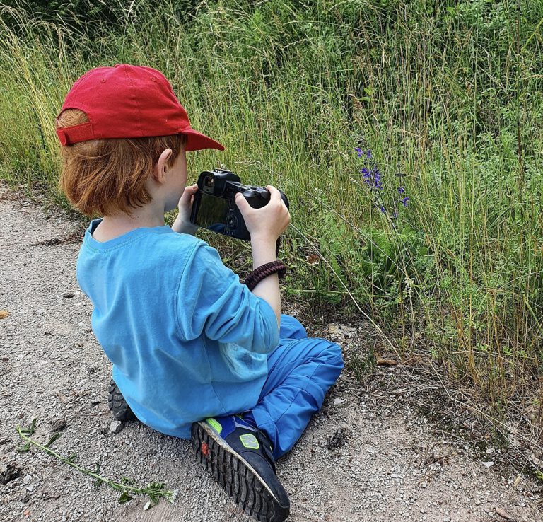 Mein Mittlerer auf dem Boden sitzend. Er hat meine Kamera in der Hand und fotografiert eine lila Blume.