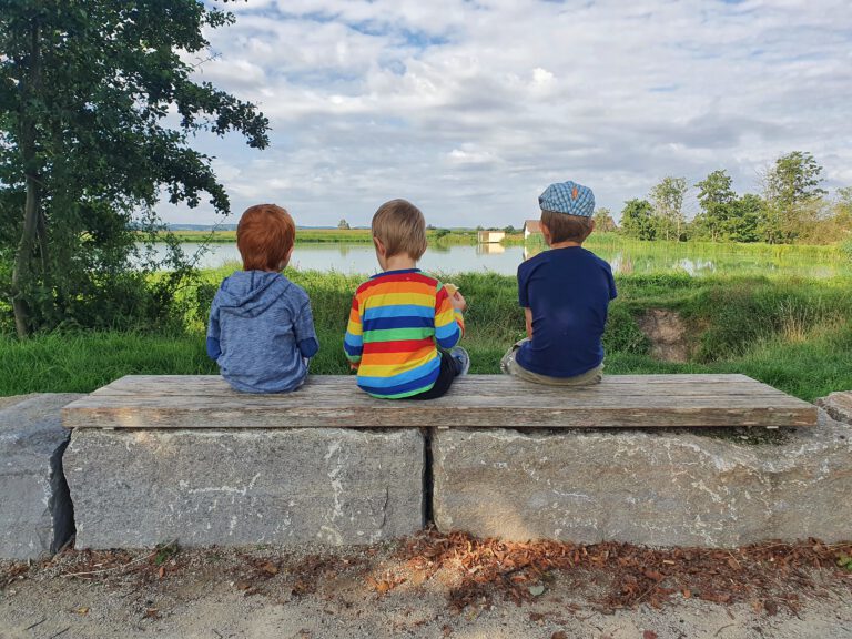 3 Kinder sitzen auf einer Steinbank und schauen in Richtung Weiher.