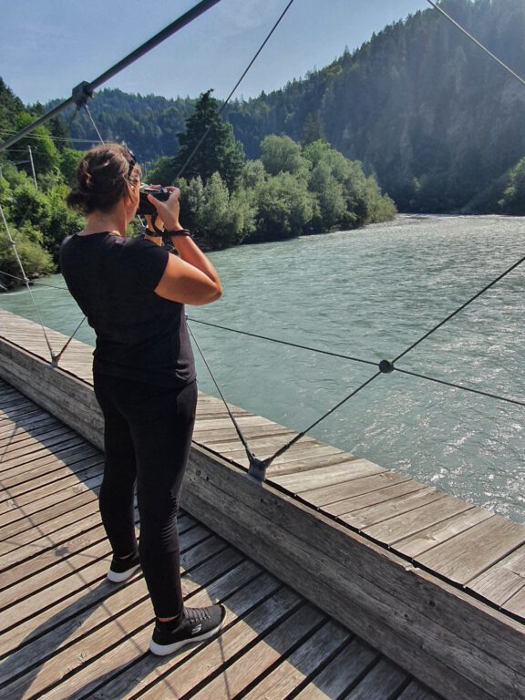 Ich in schwarz gekleidet seitlich von hinten fotografiert, wie ich auf einer Holzhängebrücke stehe und den blau grünen Rhein fotografiere.