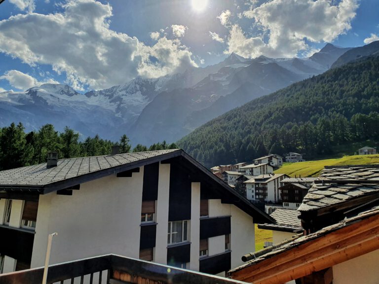 Ein Haus im Vordergrund. Dahinter grüne Nadelbäume und Berge