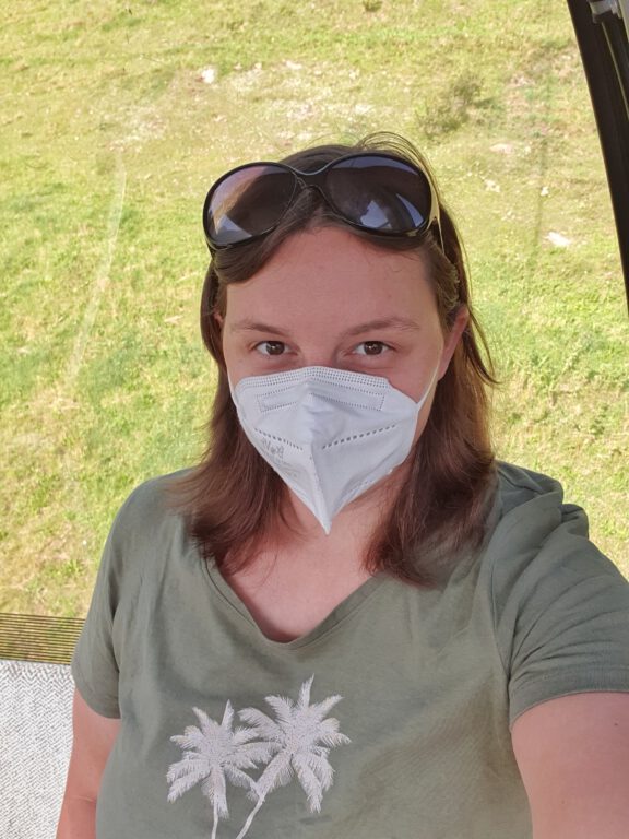 Ein Selfie von mir in einer Gondel. Ich trage eine weiße Maske und ein grünes Shirt.