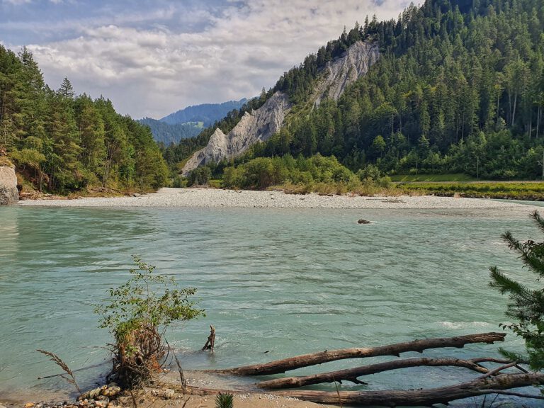 Blau grünes Gewässer umgeben von Bergen und Nadelbäumen. Etwa in der Mitte ist ein Steinstrand.