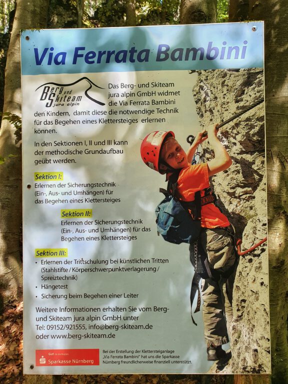 Ein Schild über den Bambini Klettersteig. Drauf zu sehen ist ein Kind, das in Ausrüstung am Felsen hängt.