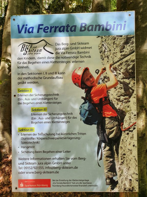 Ein Schild mit der Aufschrift "Via Ferrata Bambini" - Man sieht ein Kind in Kletterausrüstung an einem Felsen.