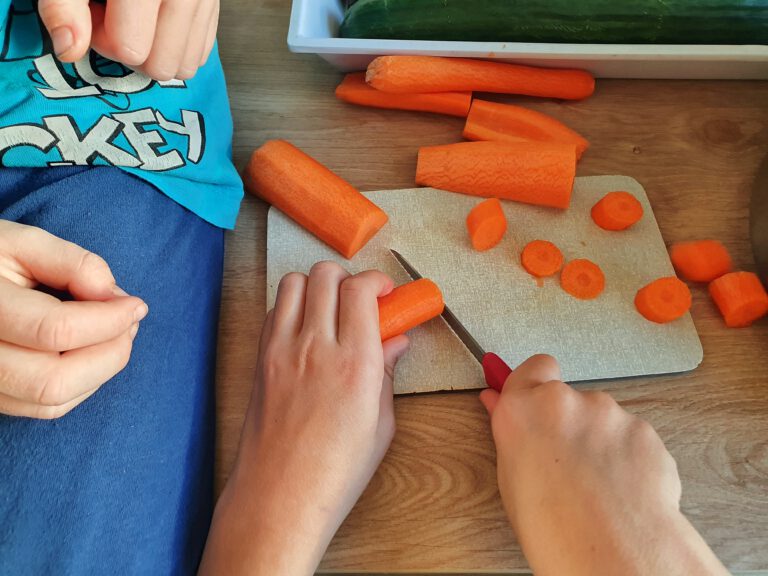 4 Kinderhände. 2 davon schneiden eine Möhre. Die anderen beiden Hände sammeln das Gemüse ein.