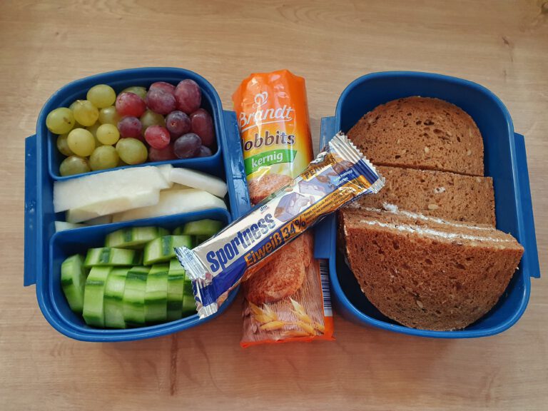 2 blaue Boxen mit Essen. In der linken sind grüne und rote Weintrauben, Kohlrabi, und Gurken. Rechts sind Brote und in der Mitte eine Packung Kekse und Eiweißriegel.