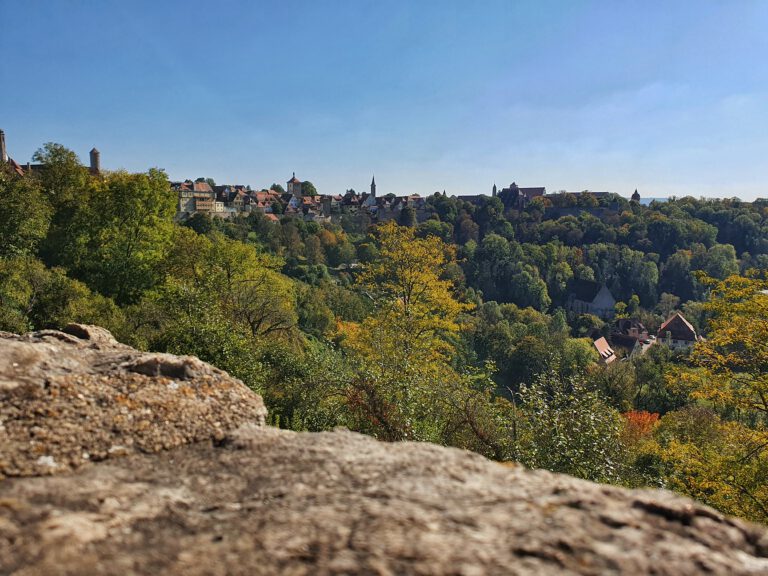 Im Vordergrund eine Burgmauer. Dahinter geht es ins Tal mit vielen Bäumen. Im Hintergrund sieht man ein paar Häuser.