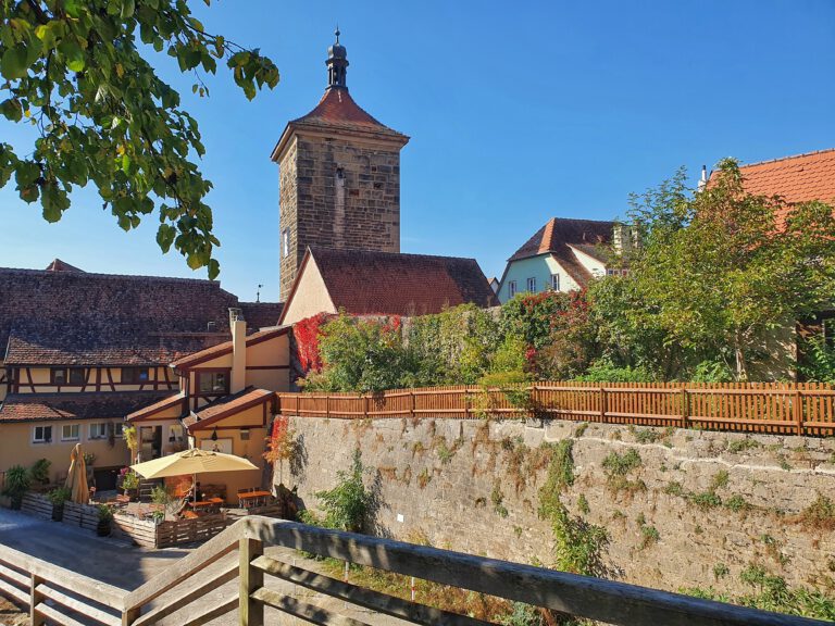 Im Vordergrund ist eine alte Burgmauer. Im Hintergrund sieht man einen großen Turm. Der Himmel ist wolkenlos blau.