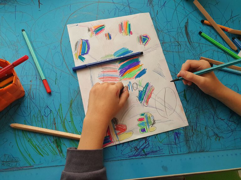 Kinderhände, die einen Stift halten und bunt auf ein Papier malen.