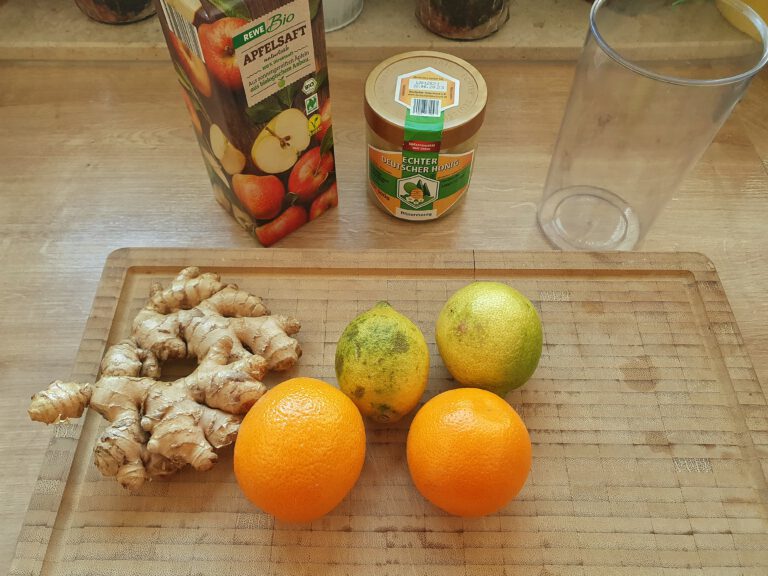 Ein Holzbrett mit Ingwer, Orangen und Zitronen. Dahinter steht Apfelsaft und Honig. 