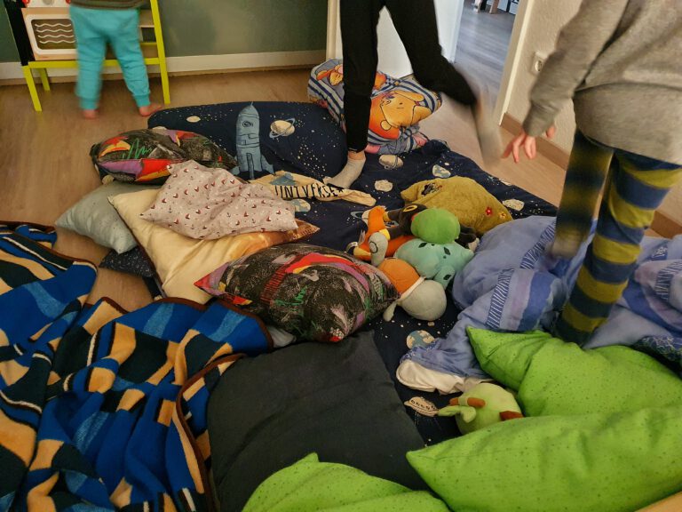 Viele Decken und Kissen auf dem Boden. Drei Kinder springen drauf herum.