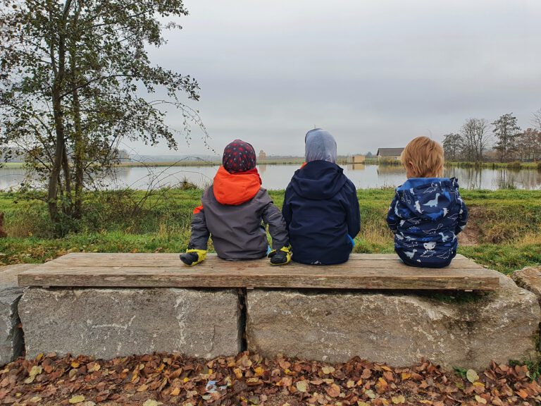 3 Kinder sitzen auf einer Bank und schauen auf einen Weiher. 