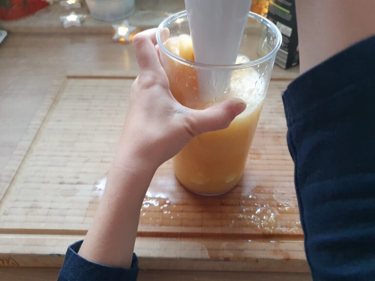 Kinderhände die einen Mixer halten und eine Orange Flüssigkeit pürieren. 