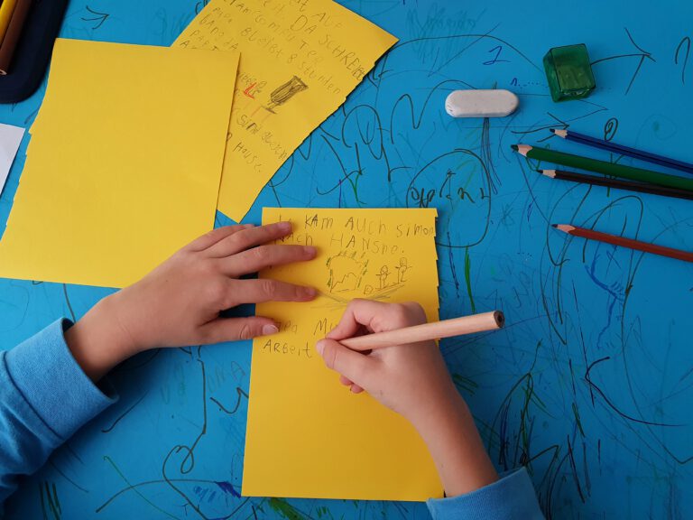 Zwei Kinderhände die einen Stift und gelben Zettel halten. Auf dem Tisch liegt eine blaue Unterlage.