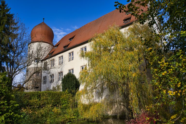 Ein großes Gebäude mit einem runden Turm an der linken Seite und einem roten Dach. Vor dem Schloß ist eine Trauerweide und weitere Büsche.