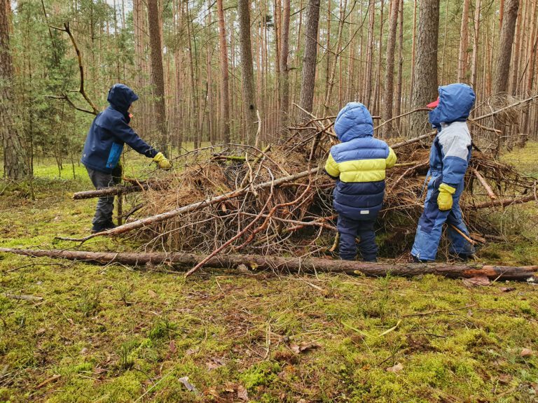 3 Kinder bauen aus Stöchen, Moos und herumliegenden Baumstämmen einen Unterschlupf für Tiere.