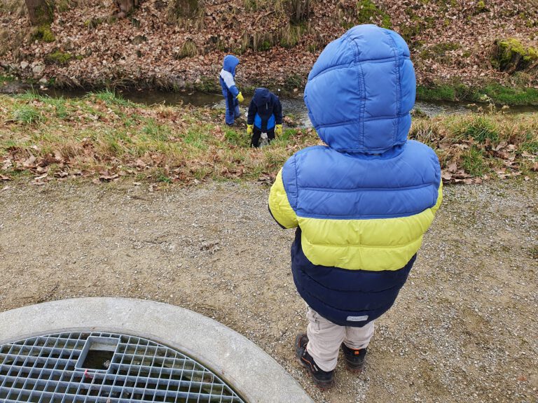 2 Kinder spielen am Bach. Ein drittes schaut zu,