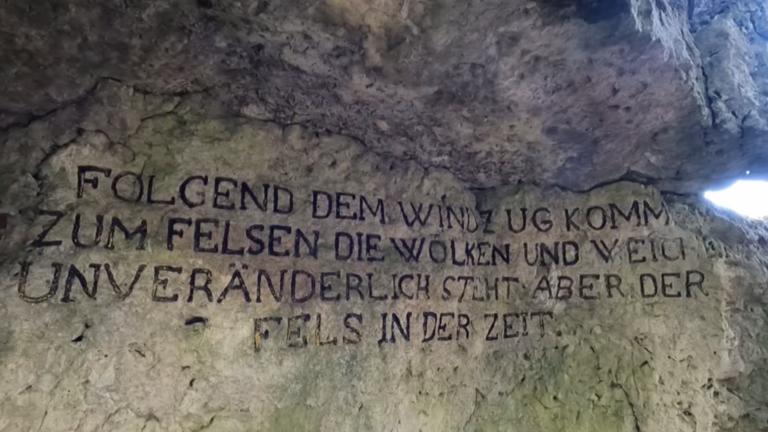 Ein in Stein gemeißelter Schriftzug: 
"Folgend dem Windzug, kommen zum Felsen die Wolken und weichen, unveränderlich steht aber der Fels in der Zeit."