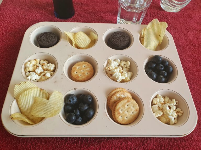 Ein Muffinblech gefüllt mit Chips, Popcorn, Keksen und Blaubeeren.