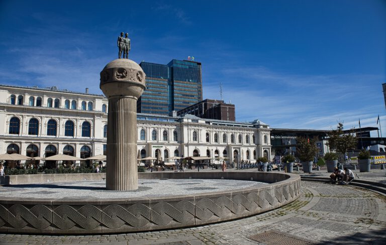 Vorplatz des Osloer Hauptbahnhofs. Ein Brunnen mit Säule und zwei Figuren oben drauf.