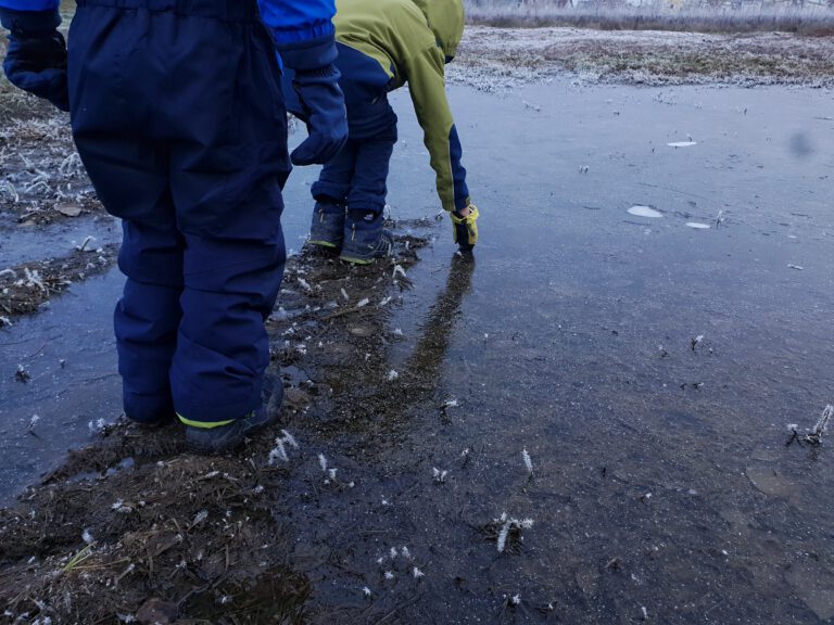 Zwei Kinder stehen am Rand einer zugefrorenen Pfütze.