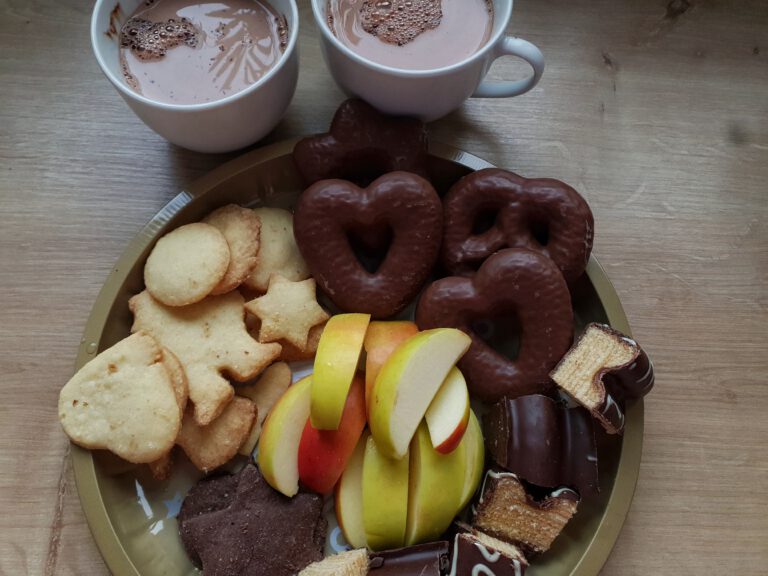 Zwei Tassen mit Kakao und einem Teller mit allerhand Süßigkeiten: Plätzchen, Lebkuchen, Äpfel und Baumkuchen.