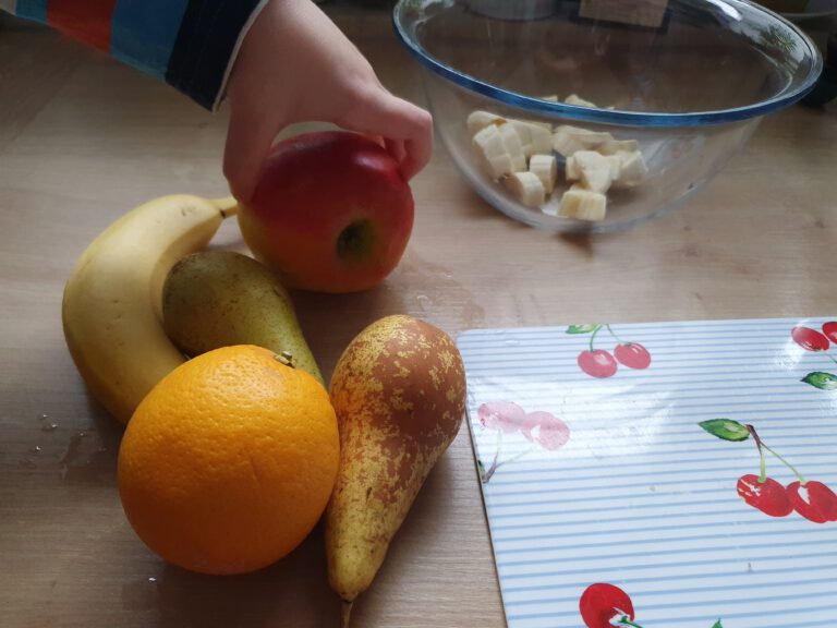 Eine Banane, Apfel, Birne, Orange und eine Schüssel. Eine Kinderhand hält den Apfel.