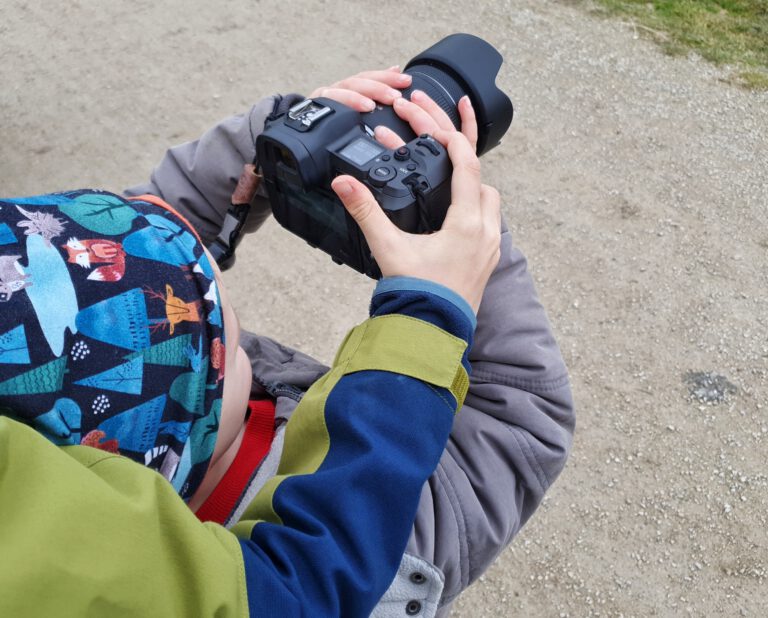 Ein kleines Kind hält eine schwarze Kamera fest. Ein größ0eres Kind steht dahinter und hilft.