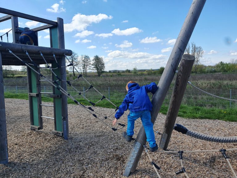 Ein Kind in blau gekleidet klettert auf einem Gerüst auf einem Spielplatz.