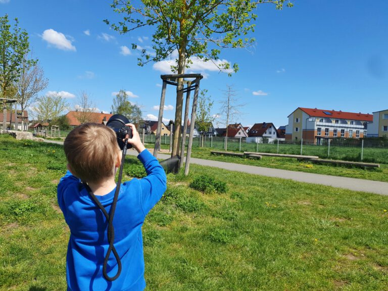 Ein kleines Kind mit blauen Shirt und einer schwarzen Kamera in der Hand. Er fotografiert einen Baum.