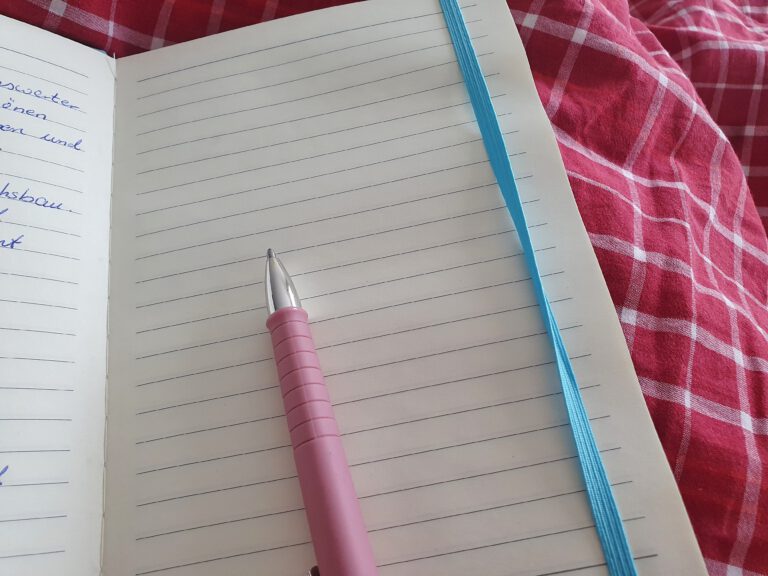 Ein Notizbuch mit blauen Band und einem rosa Kugelschreiber. Die Seite ist noch leer.