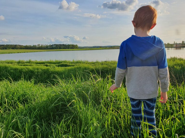 Ein Kind mit blau grauen Anziehsachen, steht im hohen grünen Gras und schaut auf einen Weiher.