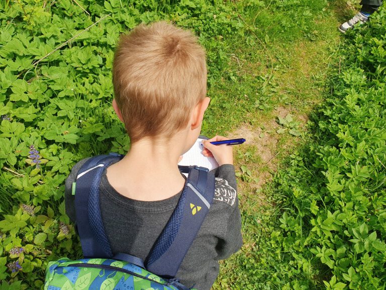 Ein Kind mit einem Rucksack, der in der Natur hockt und mit einem Stift etwas in ein Notizbuch schreibt.