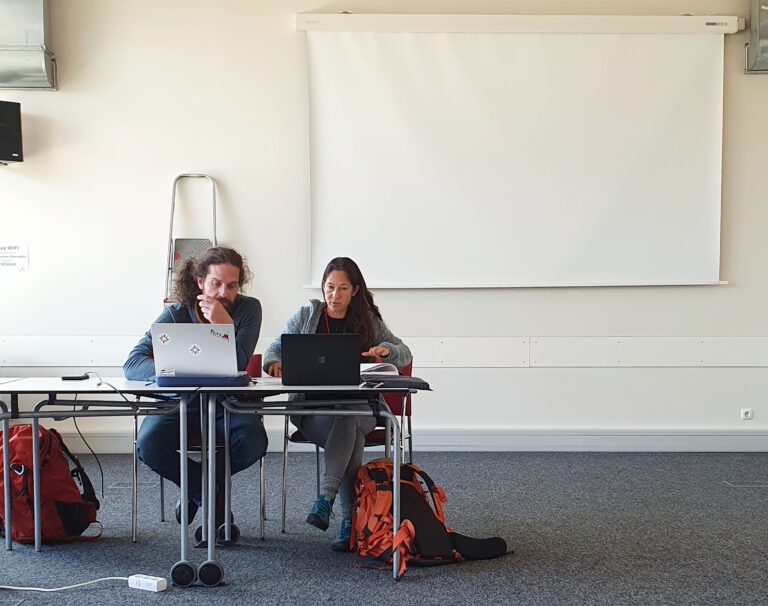 Zwei Erwachsene sitzen an einem Tisch mit Laptops. Im Hintergrund ist ein Beamer.