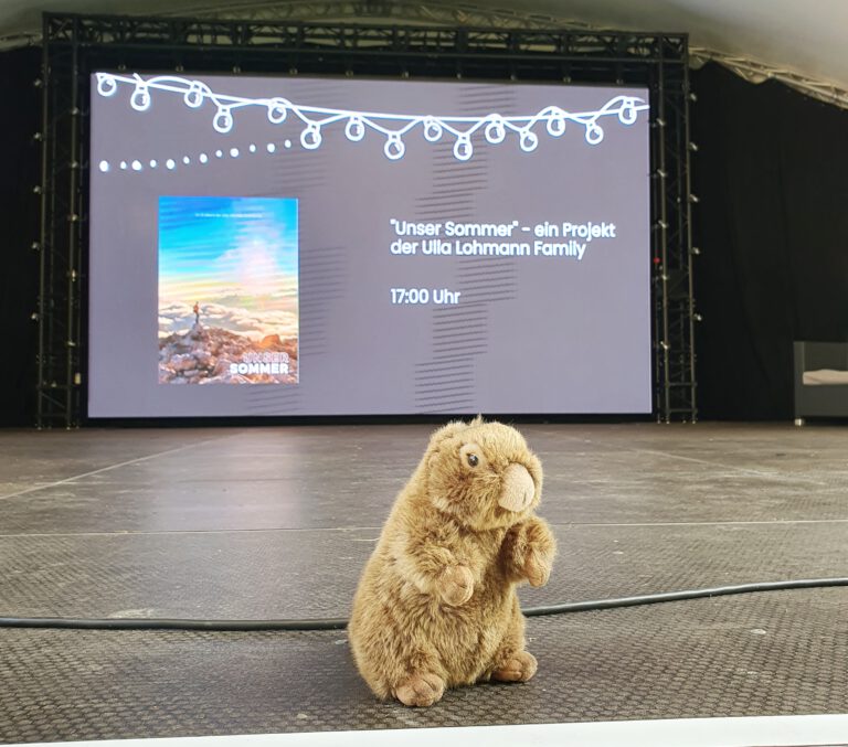 Ein stoff-Murmeltier sitzt auf einer Bühne mit Leinwand im Hintergrund.