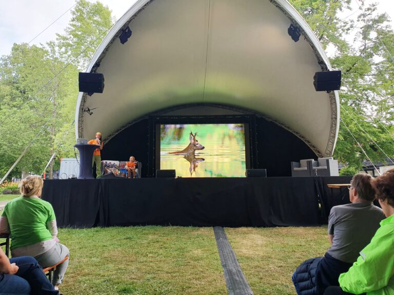 Eine Bühne im Freien mit Leinwand im Hintergrund auf dem ein Bild zu sehen ist. Im Vordergrund sitzen Personen auf einer Couch. Eine steht am Pult mit Mikrofon.