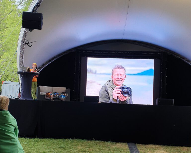 Eine Bühne im Freien mit Leinwand im Hintergrund auf dem ein Bild zu sehen ist. Im Vordergrund sitzen Personen auf einer Couch. Eine steht am Pult mit Mikrofon.