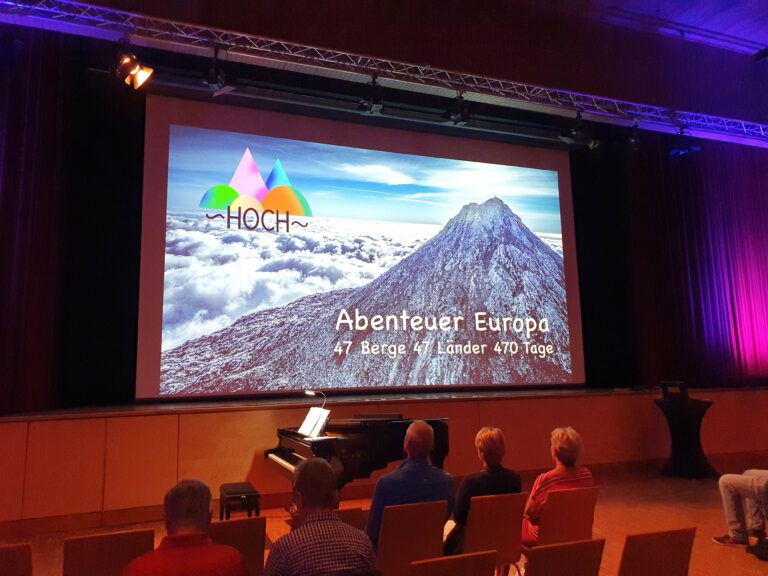 Eine Bühne in einem Saal. Im Hintergrund eine Leinwand auf der steht "Abenteuer Europa 47 Berge 47 Berge 470 Tage"