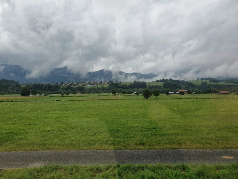 Eine weite grüne Wiese. Der Himmel ist grau mit dicken Wolken. Dahinter erahnt man die Berge.