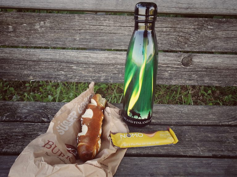 Eine Laugenstange, ein Schokoriegel und eine Trinkflasche auf einer Bank.