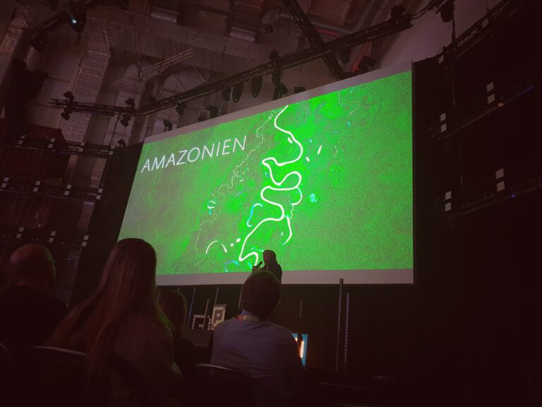 Michael Martin auf der Bühne. Im Hintergrund eine große Leinwand mit einem in grün gehaltenen Bild. Drauf steht "Amazonien".