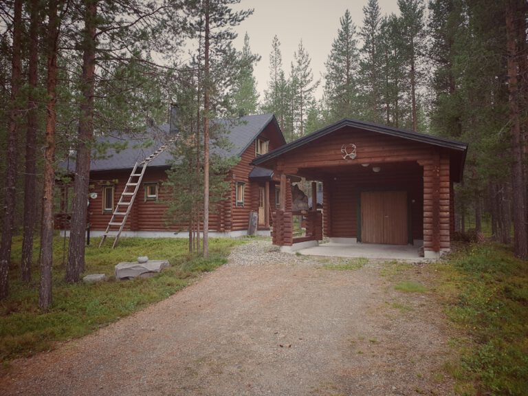 Rechts ein kleiner Unterstand aus Holz in dem ein Auto parken kann. Links  eine große Holzhütte in finnisch Lappland.