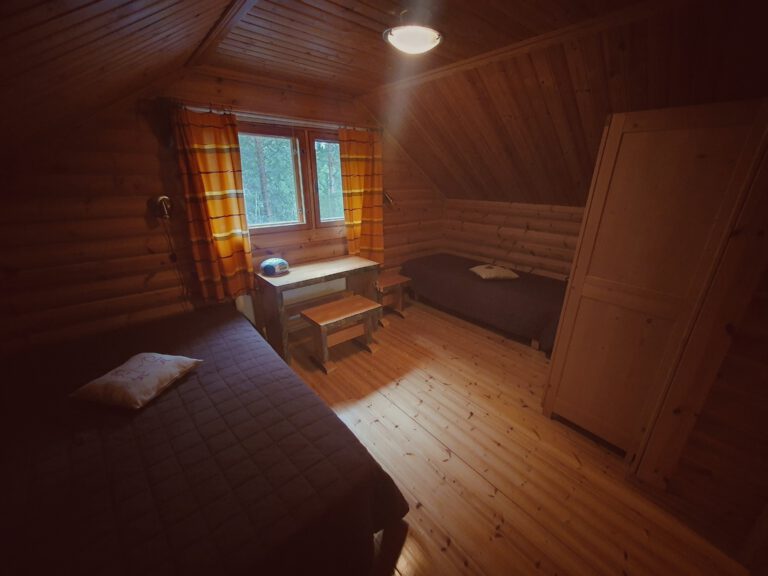 Ein Zimmer voll ausgekleidet mit Holz. Rechts und links jeweils ein Bett. Am Fenster steht ein Schreibtisch mit Stuhl davor. In der Ecke rechts ein Schrank.