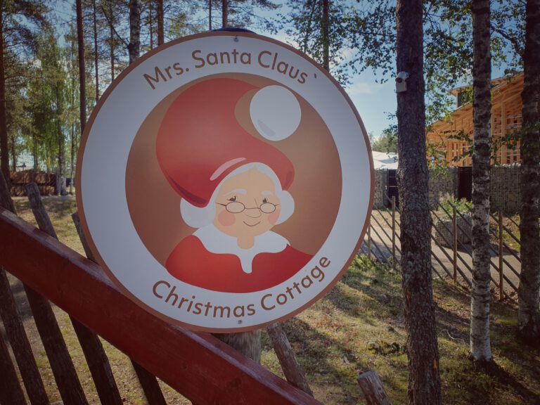 Ein rundes Schild mit einer weiblichen Figur drauf. Sie trägt eine Brille, hat rote Bäckchen und eine rot weiße Mütze auf. Drüber steht "Mrs Santa Claus"