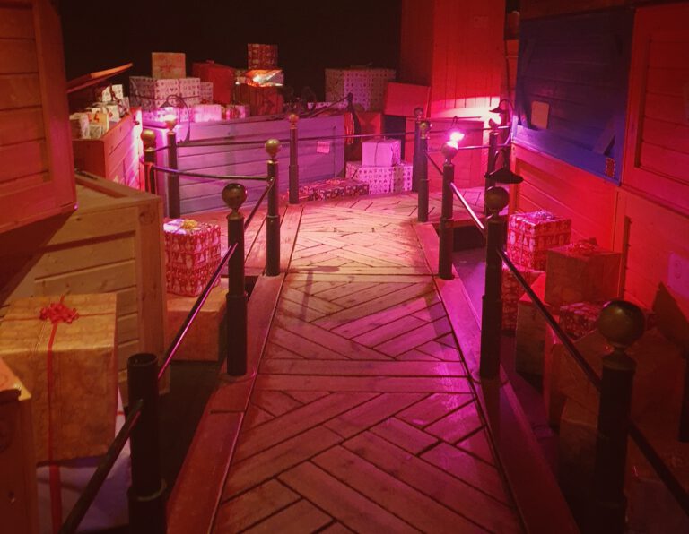 Ein Gang mit rotem und pinken Licht. Rechts und links sind viel Kisten und Geschenke aufgestellt.