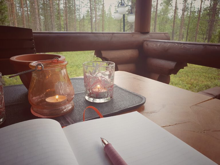 Zwei Teelichter brennen auf einer Holzterrasse. Davor liegt ein leeres Notizbuch mit Stift.