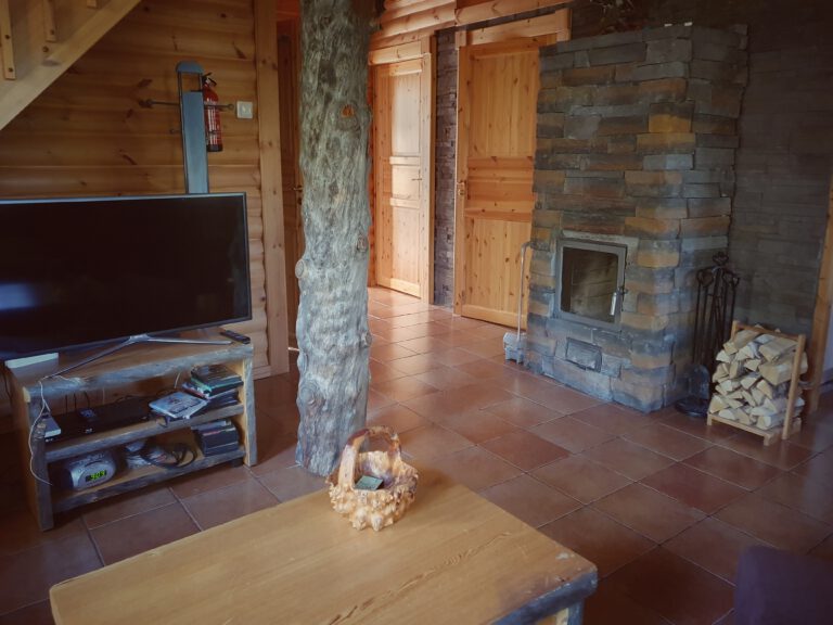Ein Kamin aus Stein. mit Holz daneben. Links ein Fernseher. Der Raum besteht fast nur aus Holz und einem Holzbalken in der Mitte.