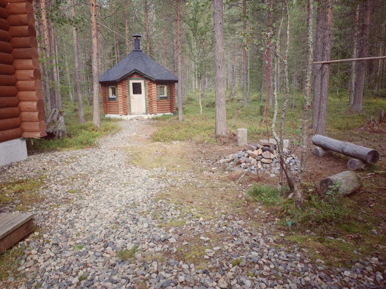 Eine Lagerfeuerstelle aus Stein mit Holzbalken drumherum. Im Hintergrund eine Runde Holzhütte mit Kamin.